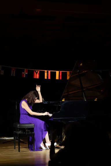 Une pianiste en robe violette joue du piano sur scène, avec des drapeaux en arrière-plan.